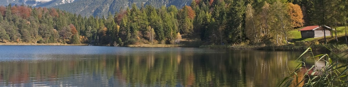 Un lac paisible où se reflètent les arbres et les montagnes, entouré d’un feuillage automnal et bordé d’une petite cabane.