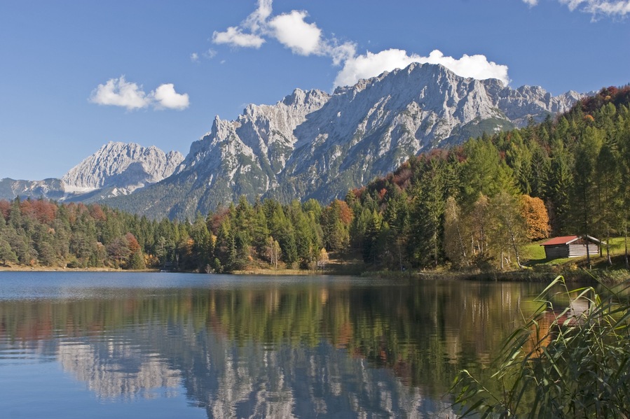 Un lago tranquilo con reflejos de árboles y montañas, rodeado de follaje otoñal y una cabaña pequeña.