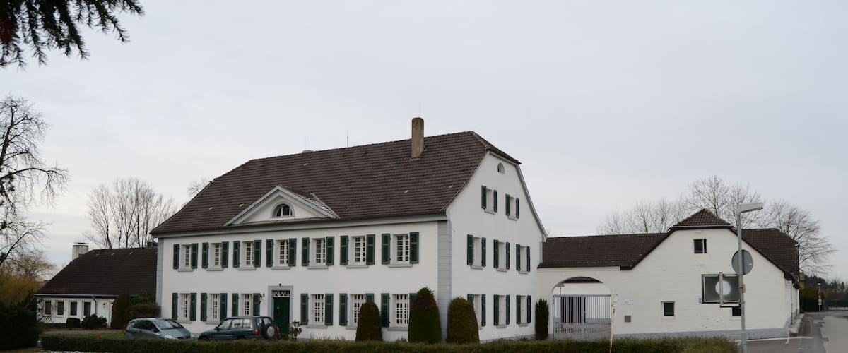 A large white house with green shutters, a garage, and a driveway with parked cars.