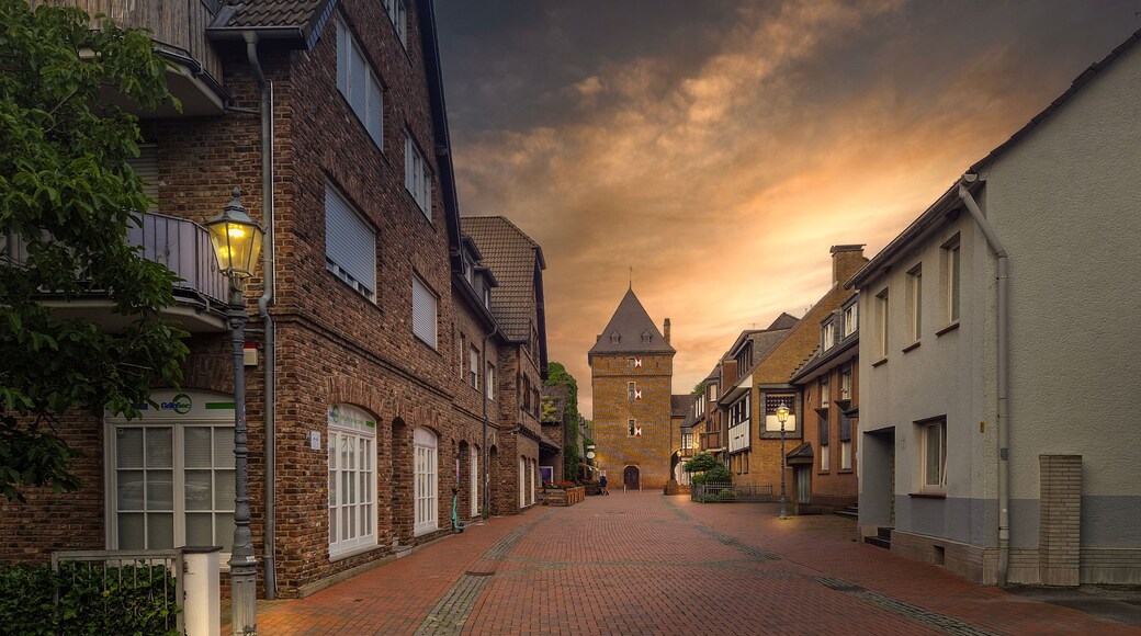 Een geplaveid straatje omringd door historische gebouwen, met een toren in de verte en een zonsonderganglucht.