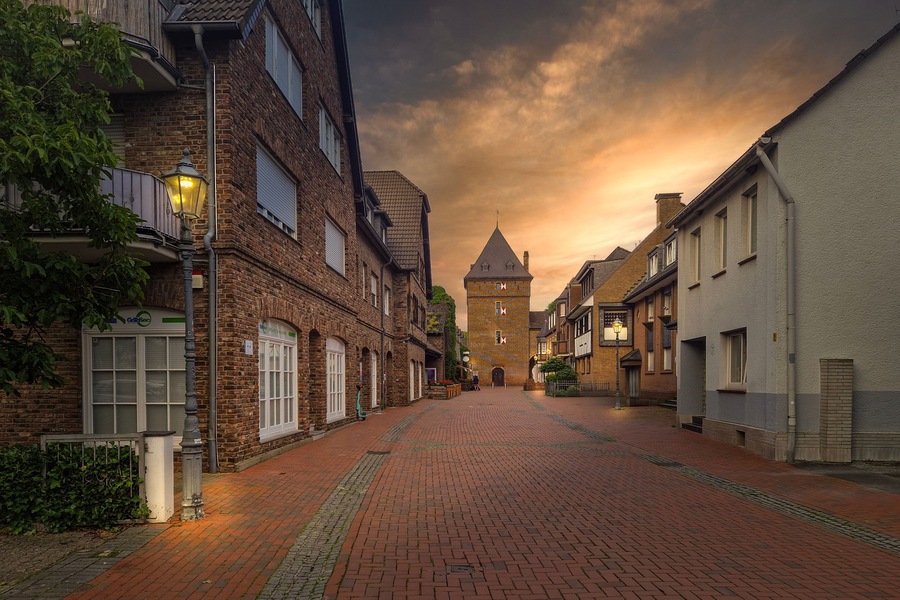 A cobblestone street lined with historic buildings, a tower in the distance, and a sunset sky.