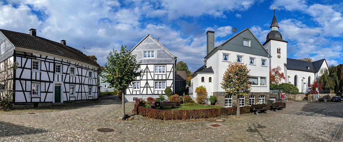 A cobblestone square with traditional half-timbered houses, a church with a steeple, and a sign for a restaurant.
