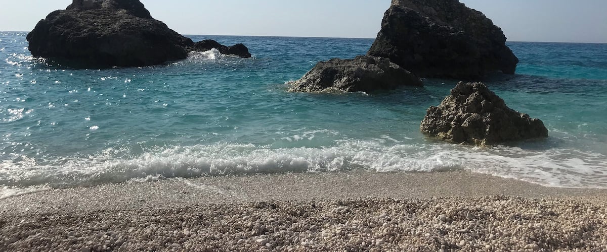 A beach with pebbly shore, clear blue sea, and rocky formations.