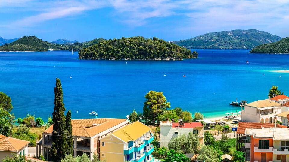 A coastal town with Mediterranean architecture, a clear blue sea, and lush green hills in the background.