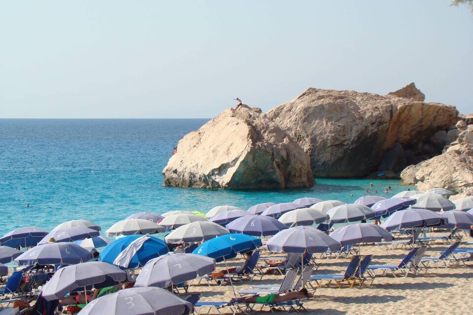 A beach with numerous umbrellas and sun loungers, rocky formations in the background, and clear blue water.