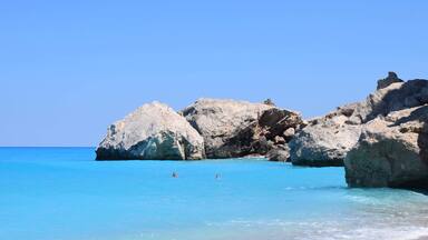 A clear blue sea with rocky formations and two people swimming.