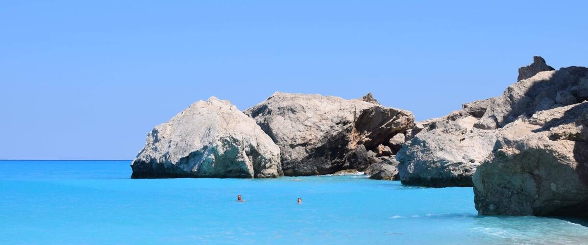 A clear blue sea with rocky formations and two people swimming.