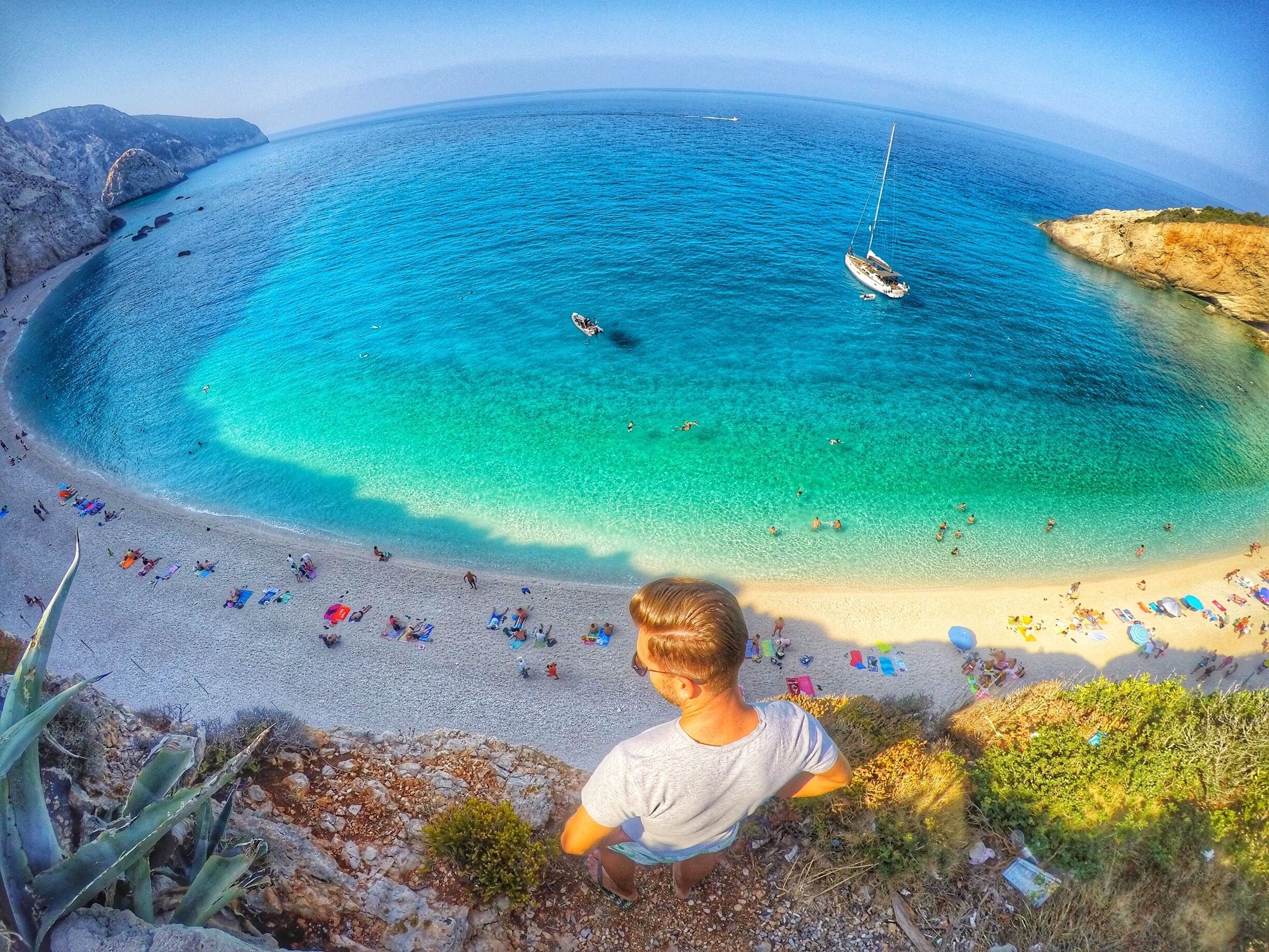 A coastal beach with clear turquoise waters, a sailboat, and people enjoying the shore.