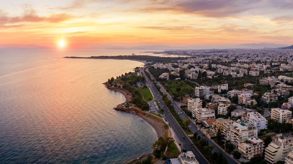 A coastal cityscape at sunset with a clear view of the sea, a prominent road along the coast, and dense urban buildings.