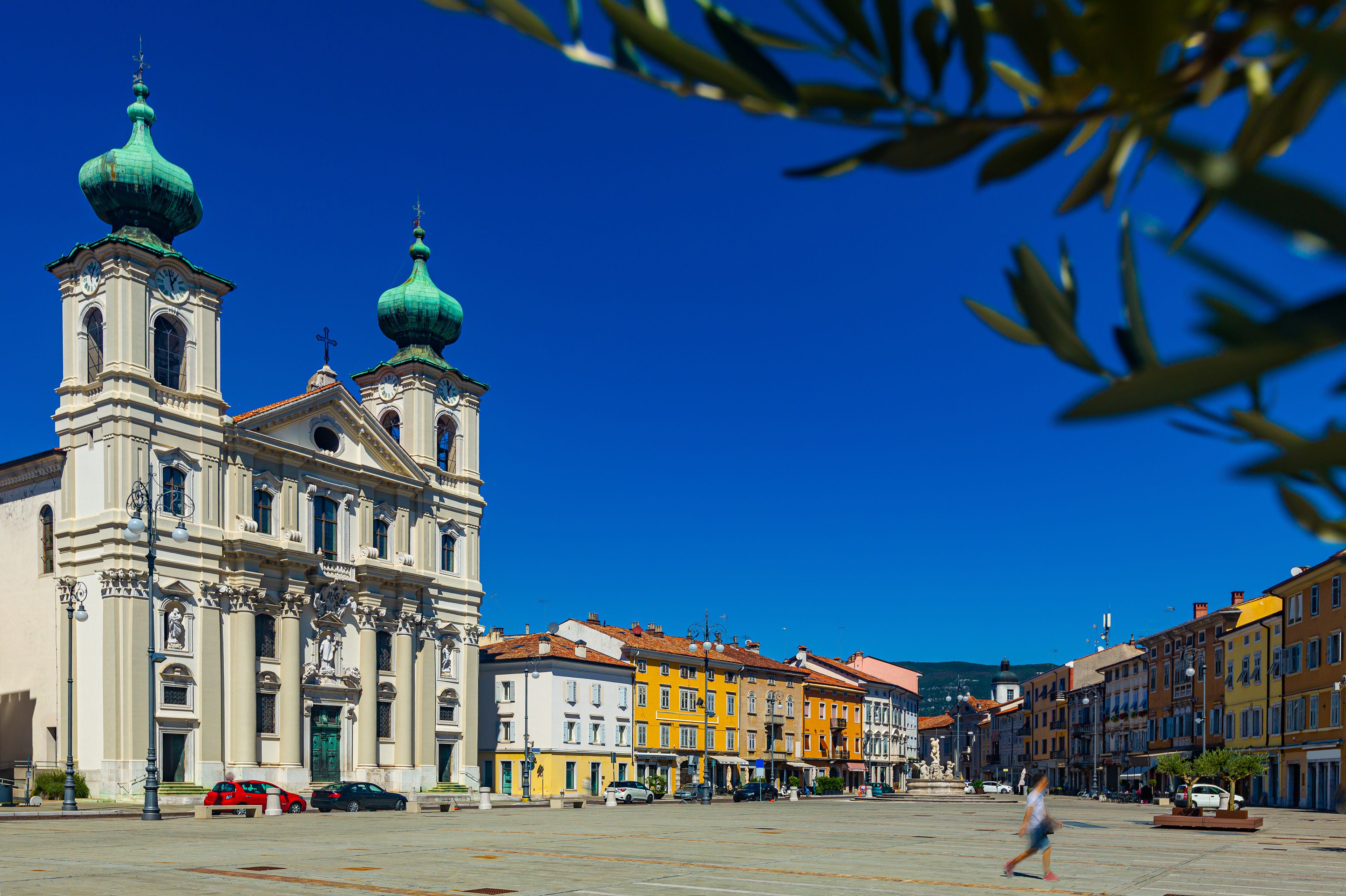 A historic church with two green domes and a clock tower, surrounded by colorful buildings and a clear blue sky.