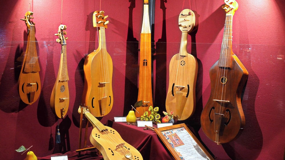 Une exposition de divers instruments de musique à cordes, notamment des guitares et des luths, sur un fond rouge, avec un document encadré et des objets décoratifs.