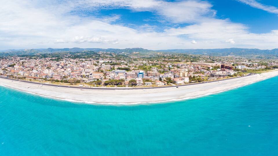 A coastal city with a long sandy beach, a pier extending into the sea, and a backdrop of mountains.