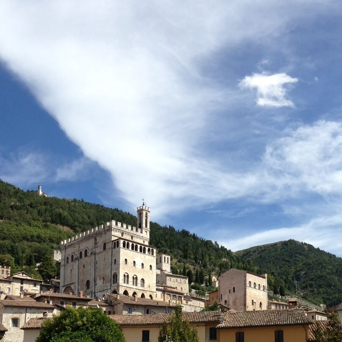 A historic stone building with a tower, surrounded by terracotta-roofed houses and lush green hills under a partly cloudy sky.