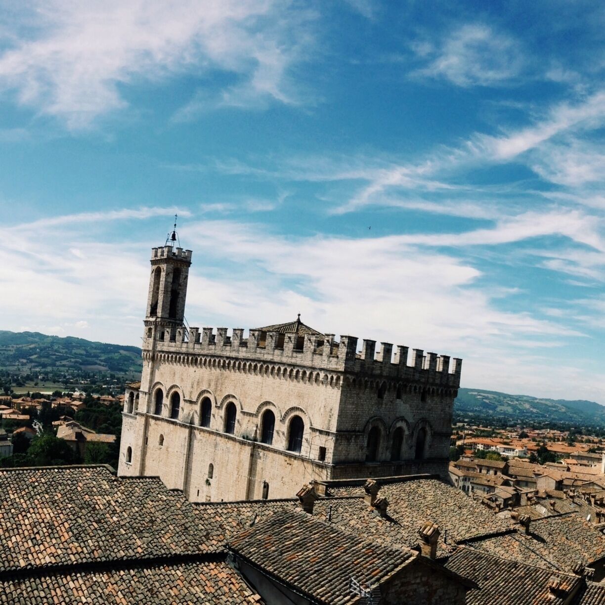 A historic stone building with a tower, surrounded by terracotta rooftops and a mountainous landscape.