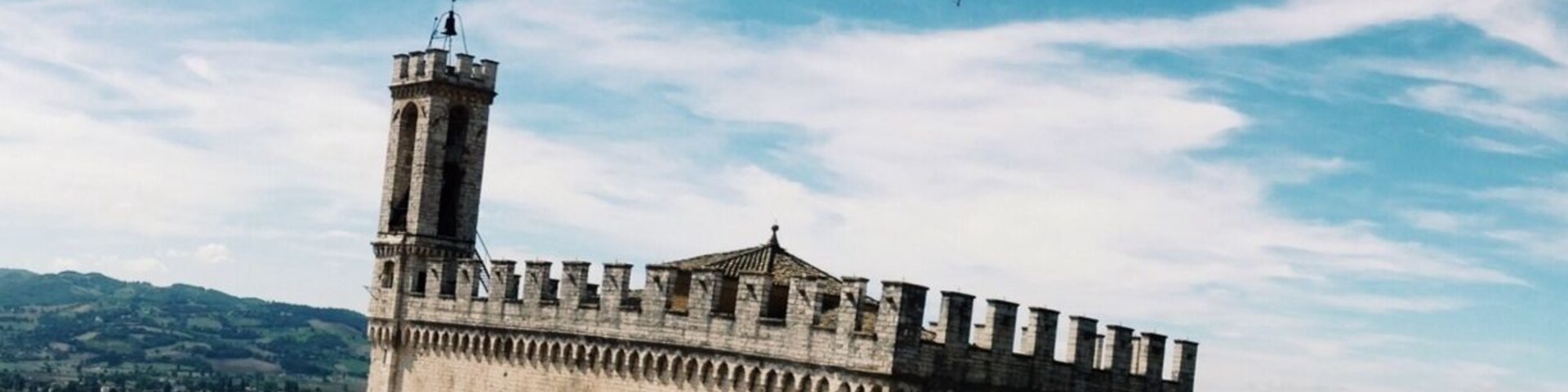 Un edificio histórico de piedra con una torre, rodeado de tejados de teja y un paisaje montañoso.