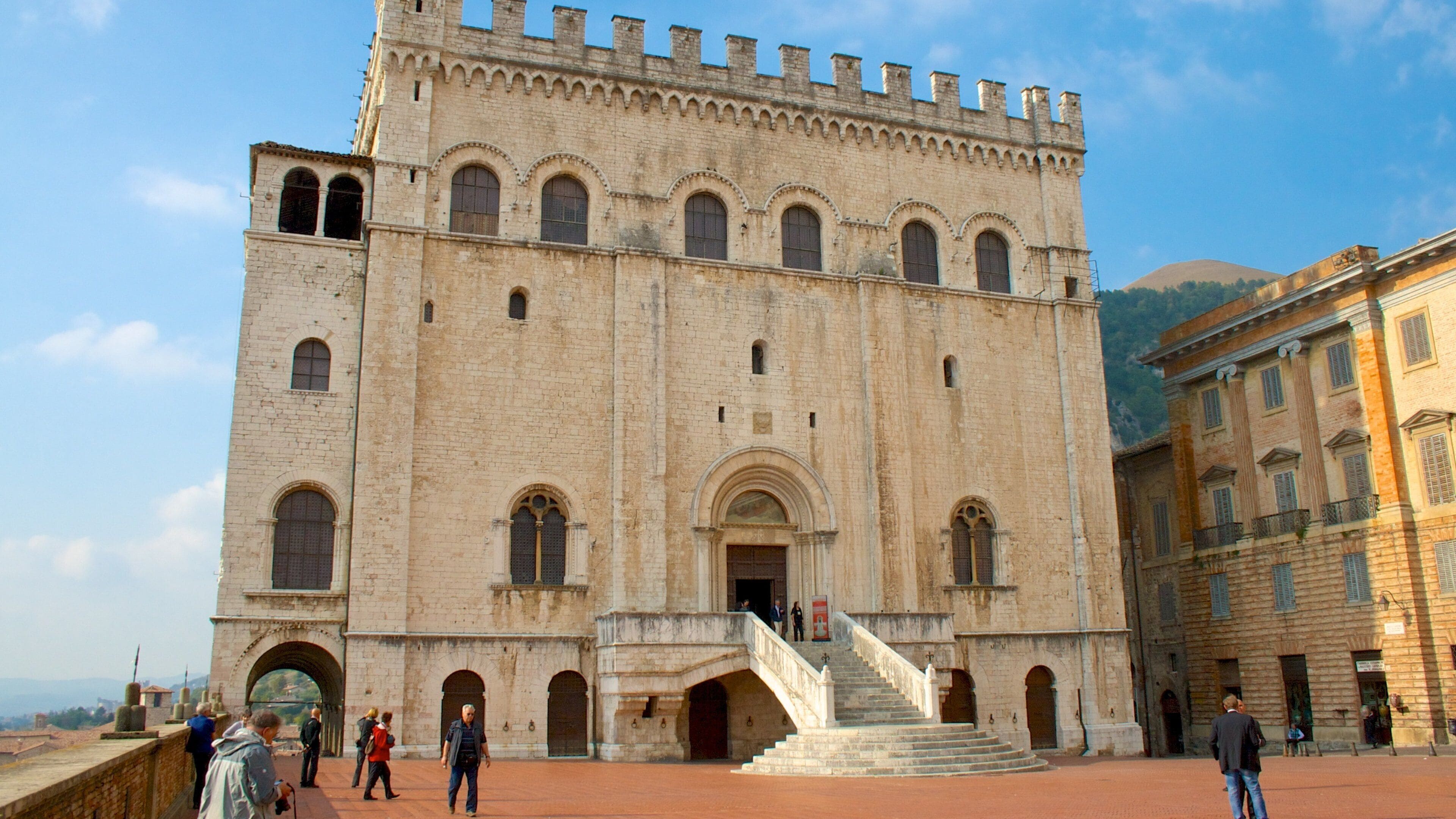 A historic building with a large arched entrance, a staircase leading up to the main entrance, and a surrounding paved area.