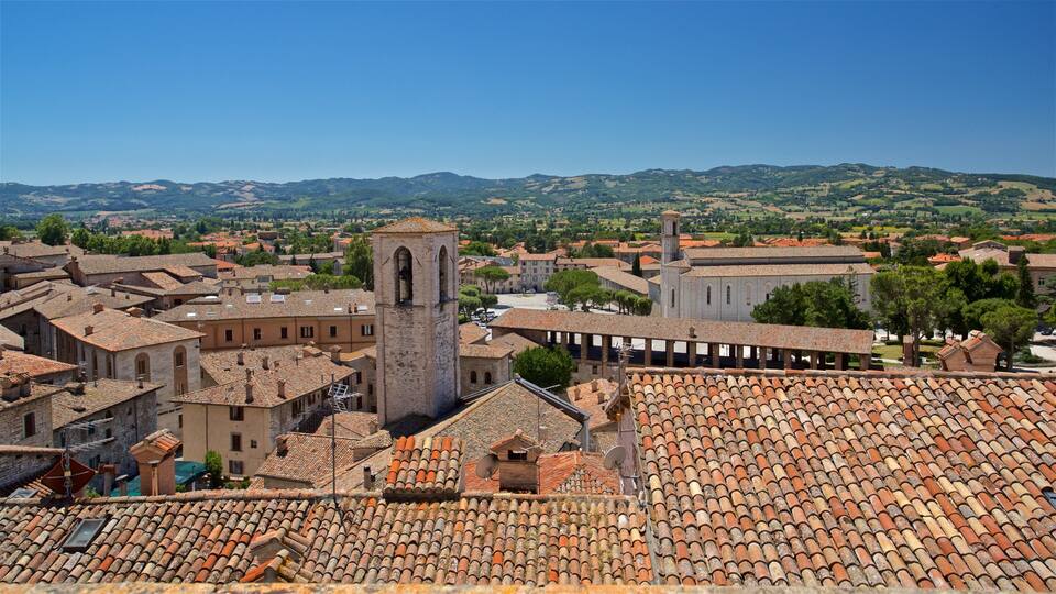 A historic town with terracotta-roofed buildings, a prominent church tower, and rolling hills in the background.