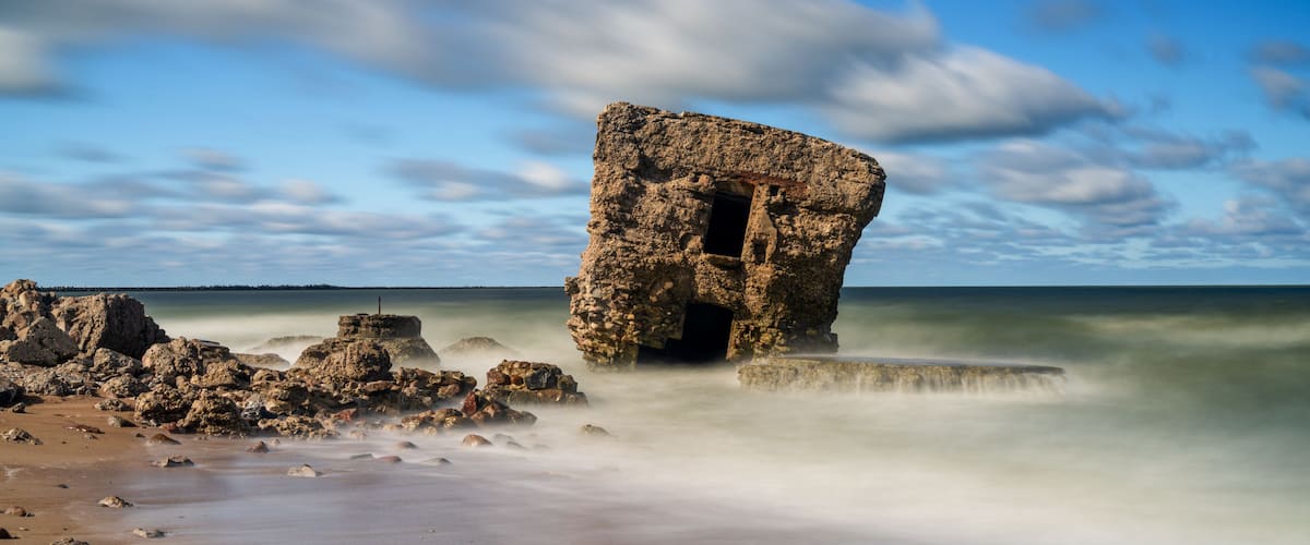 A rocky coastal landmark with a cave-like structure, surrounded by a sandy beach and calm waters.