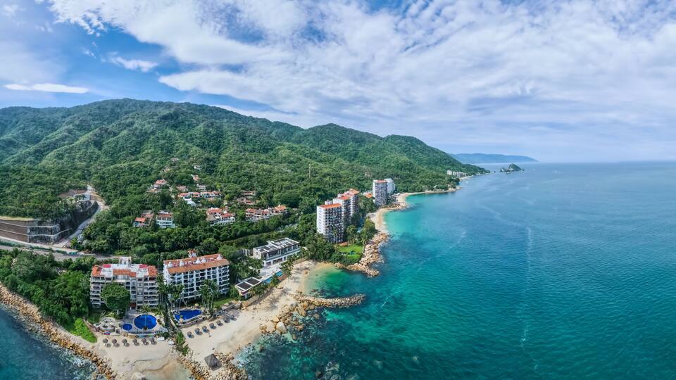 A coastal resort with a beach, clear turquoise waters, and a mountainous backdrop.