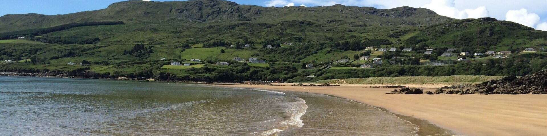 A sandy beach with gentle waves, a clear sky, and green hills in the background.