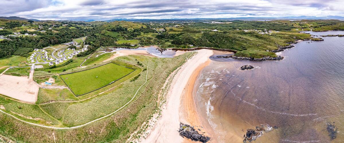 An aerial view of a coastal area with a sandy beach, green fields, and scattered houses.