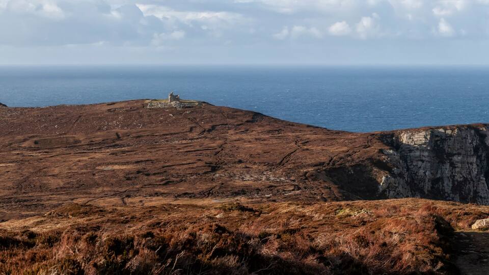 A rugged coastal landscape with a prominent cliff and a small structure on a hill.