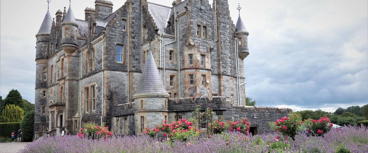 A large, historic stone castle with turrets and a well-maintained garden with purple flowers.