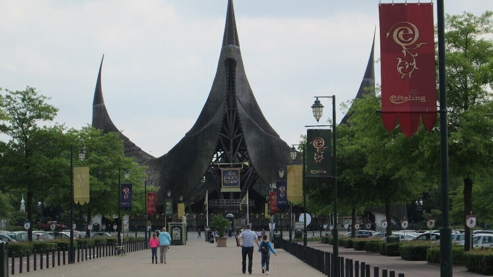 A wide pedestrian walkway with a distinctive modern structure in the background, banners hanging from lamp posts, and trees lining the path.