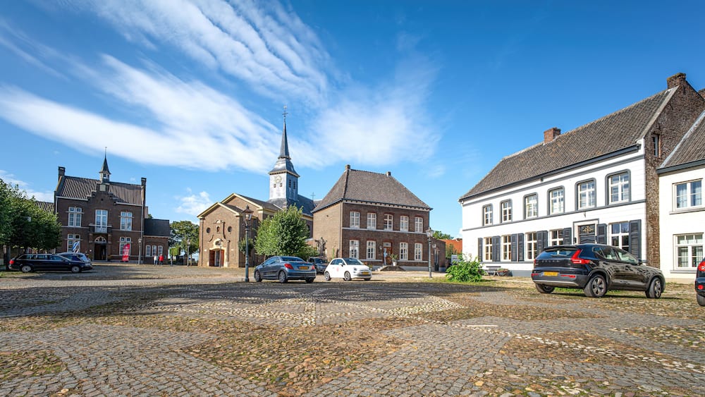 Eine Kopfsteinpflasterstraße mit historischen Gebäuden, einem Kirchturm und parkenden Autos.