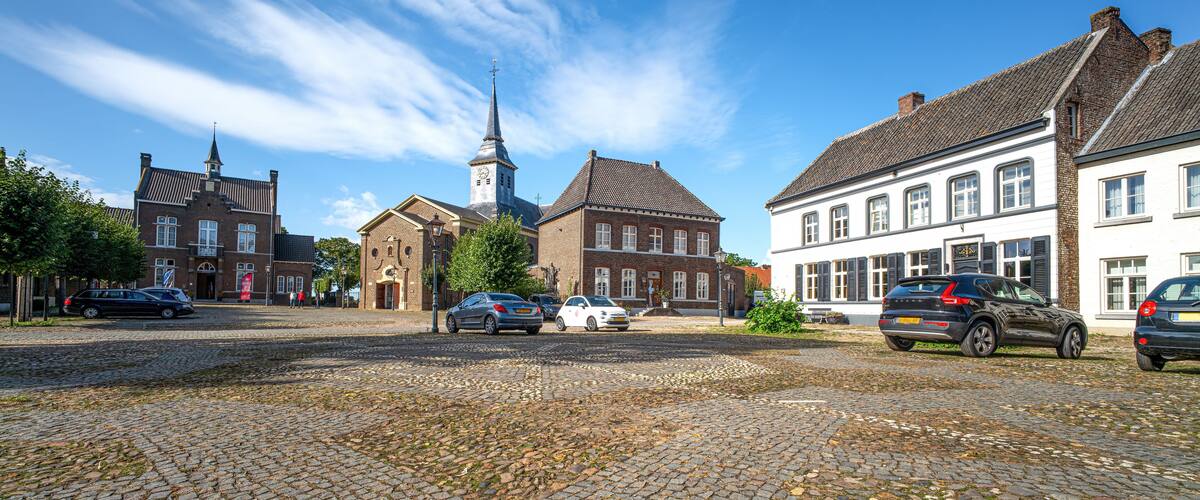 A cobblestone street with historic buildings, a church tower, and parked cars.