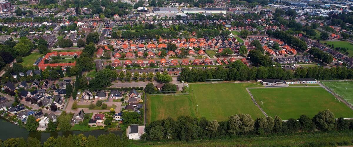 An aerial view of a town with red-roofed houses, a large green field, and a river.