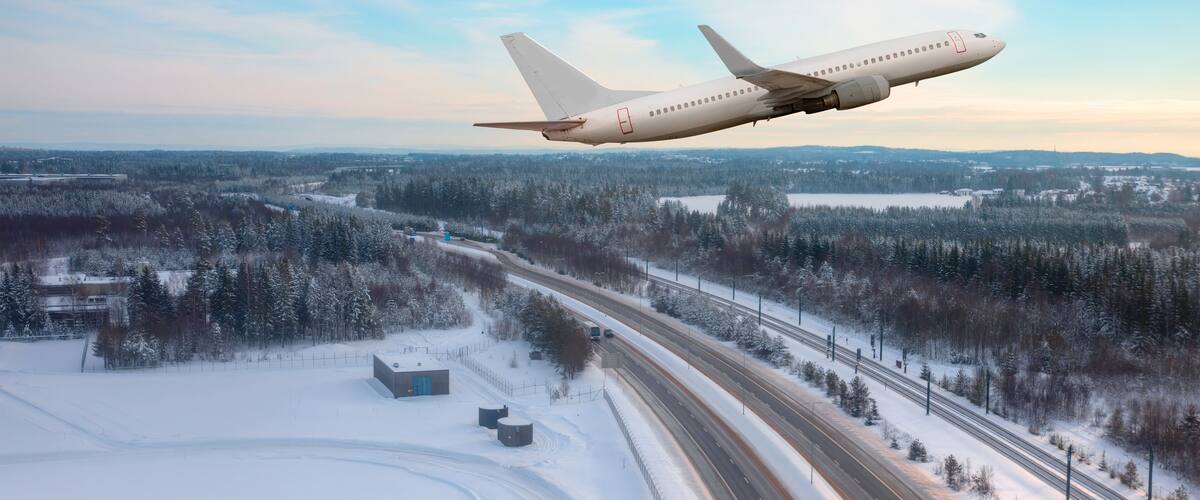 A large airplane flying over a snowy landscape with a highway and forested area.