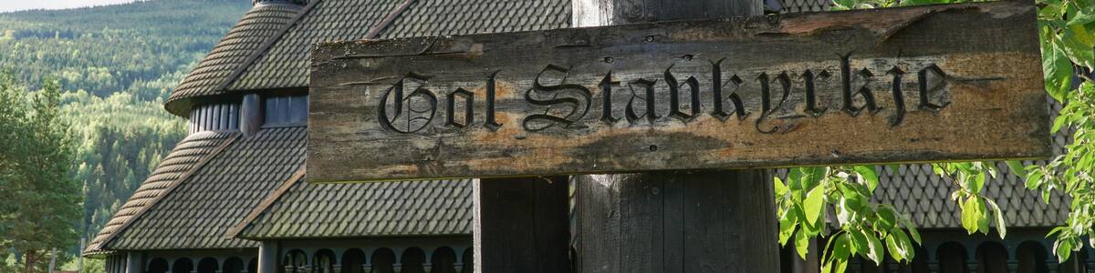 A wooden sign with 'Hol Stavnkyrk' carved on it, a traditional wooden building with a steep roof, and a forested hill in the background.