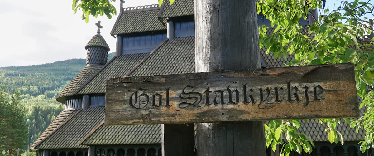 Une enseigne en bois sur laquelle est gravĂ© le nom « Hol Stavnkyrk », un bĂątiment traditionnel en bois dotĂ© dâun toit en pente et, en arriĂšre-plan, une colline boisĂ©e.