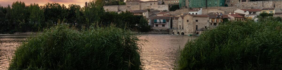 Photograph of Zamora, highlighting the Cathedral, seen from the other side of the Duero River at dusk, with the setting sun bathing the scene in golden and orange hues. Zamora, Spain