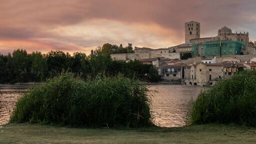 Photograph of Zamora, highlighting the Cathedral, seen from the other side of the Duero River at dusk, with the setting sun bathing the scene in golden and orange hues. Zamora, Spain