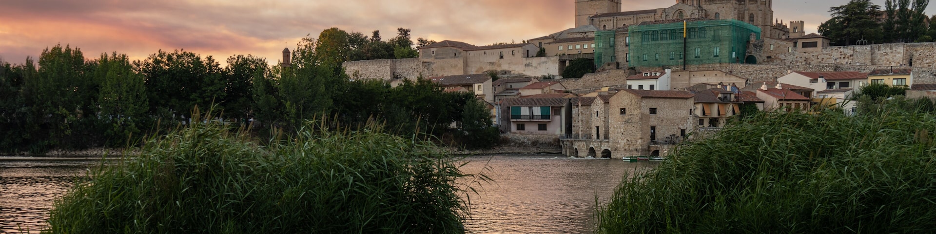 Photograph of Zamora, highlighting the Cathedral, seen from the other side of the Duero River at dusk, with the setting sun bathing the scene in golden and orange hues. Zamora, Spain