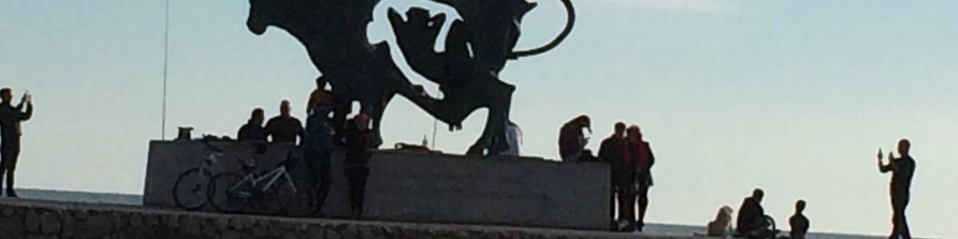 Une sculpture de taureau avec des cavaliers, placée sur une plateforme en pierre au bord de la mer.