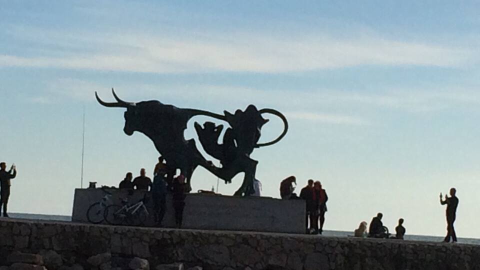 A bull sculpture with riders on a stone platform by the sea.