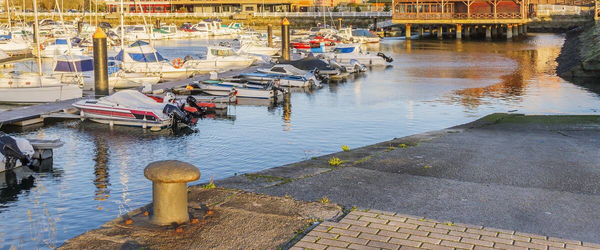 A marina with boats docked, a waterfront promenade, and buildings in the background.