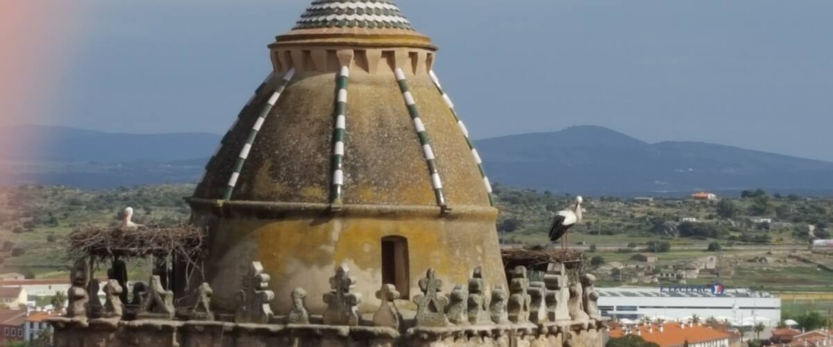 A domed building with a tiled roof, surrounded by statues and a mountainous landscape in the background.