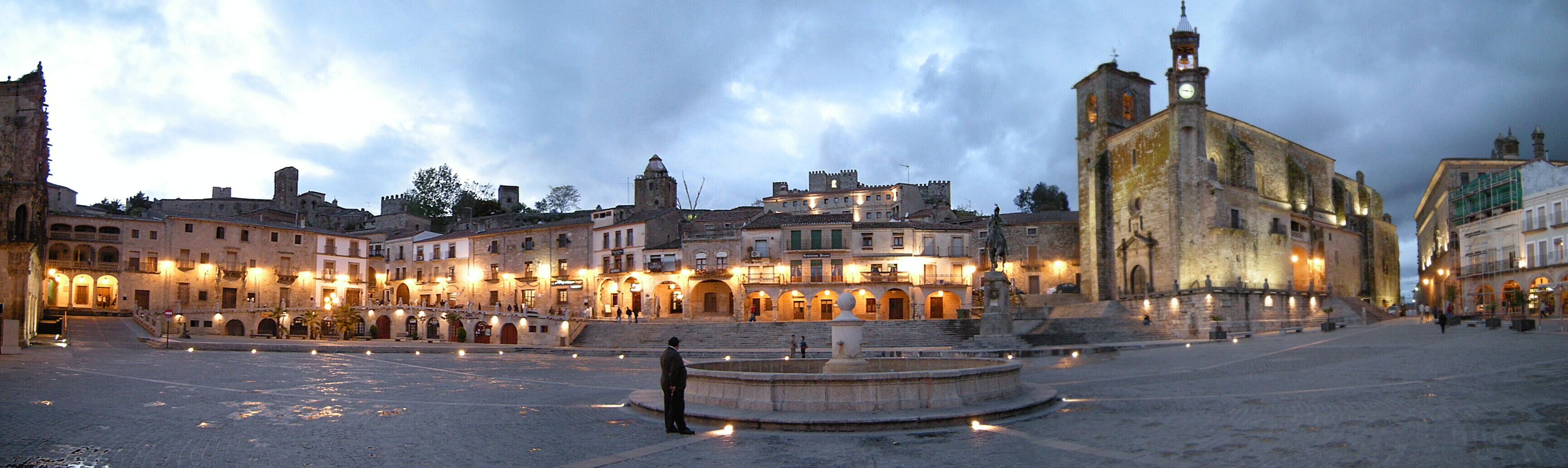 A historic square with a fountain, surrounded by old buildings and a prominent church with two towers.