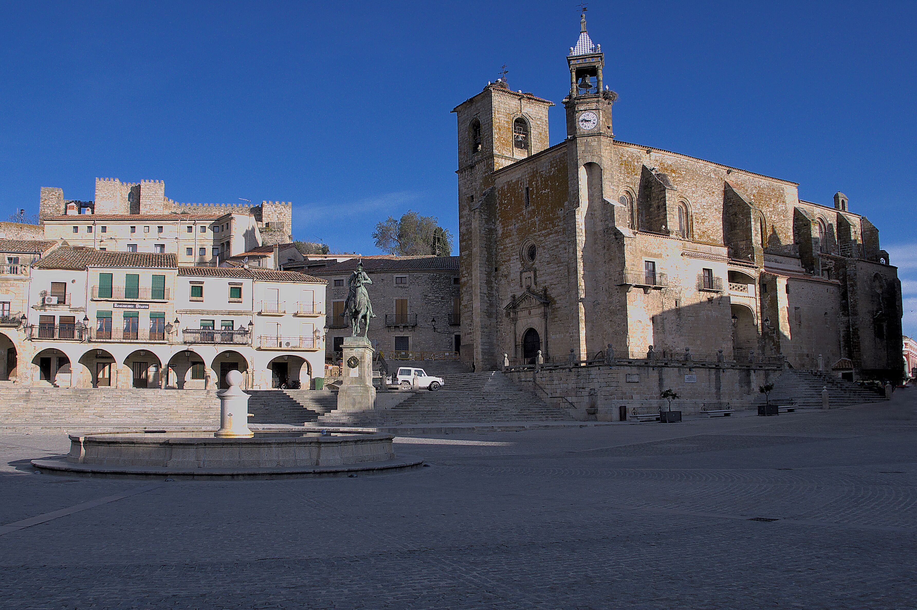 A historic square with a prominent church featuring a bell tower, a statue, and a fountain.
