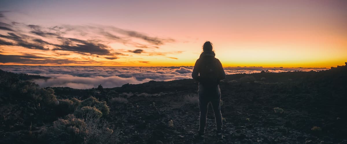 A person standing on a rocky outcrop overlooking a valley with a sunset in the background.