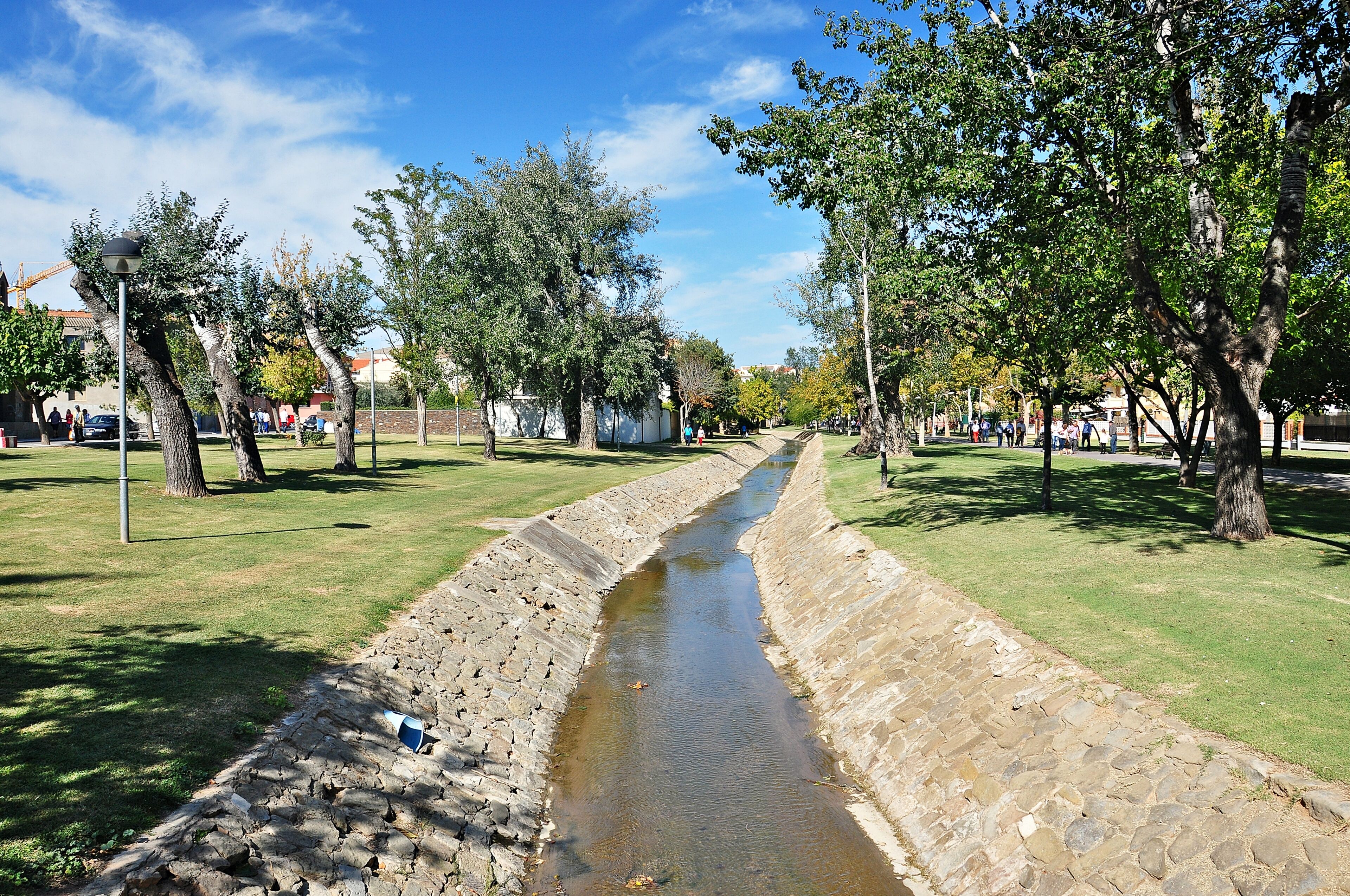 A narrow water channel flanked by trees and grassy areas, with people in the background.
