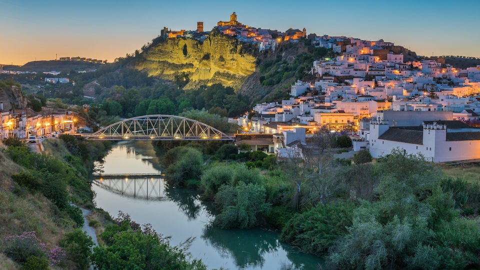 A river with a bridge, a hilltop fortress, and a densely packed town with white buildings.