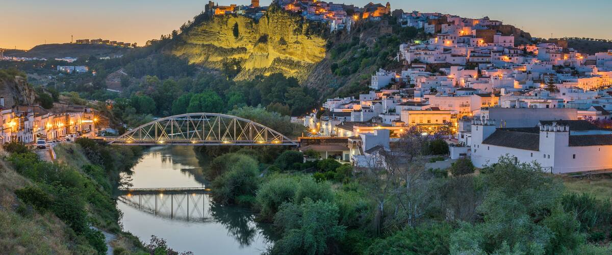 A river with a bridge, a hilltop fortress, and a densely packed town with white buildings.