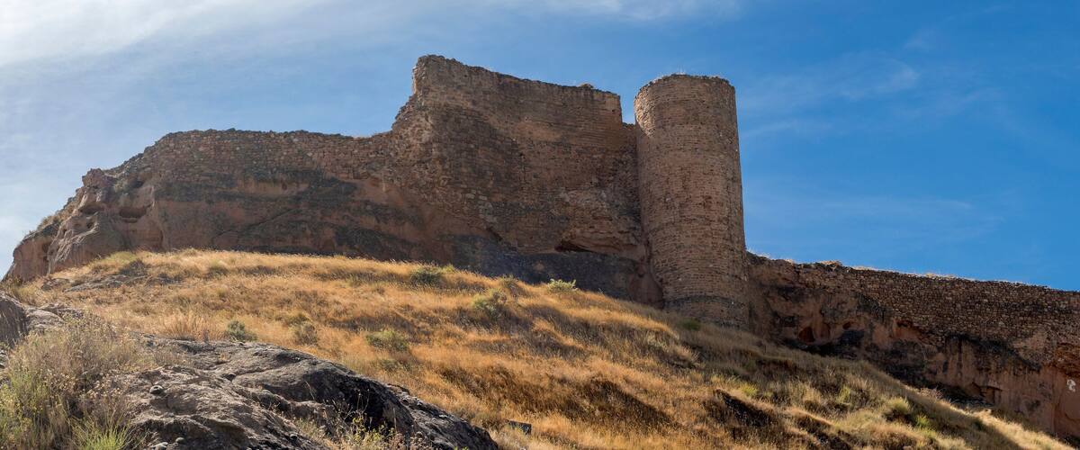 A large, ancient stone structure with a tall tower, surrounded by dry grass and rocky terrain.