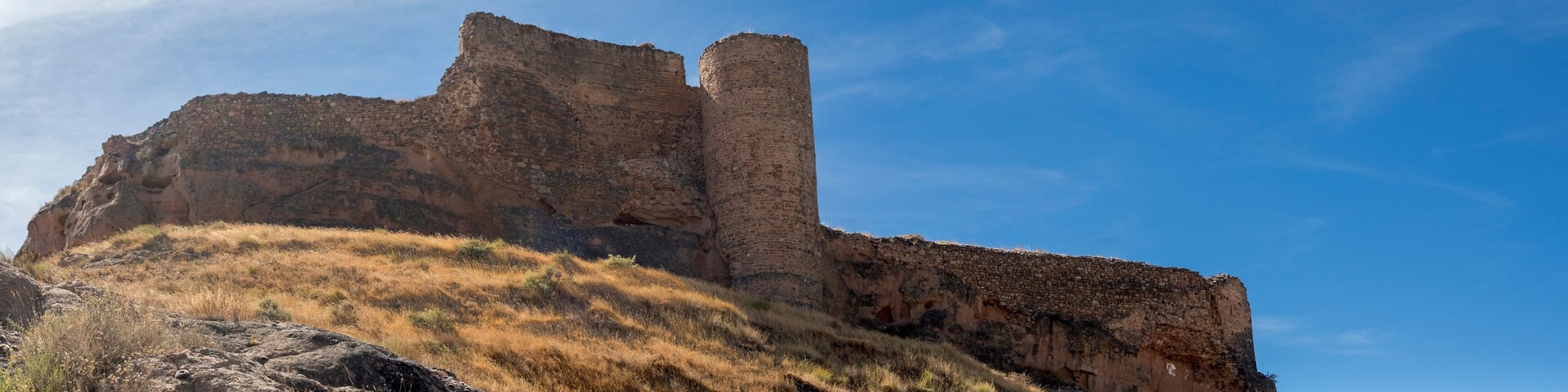 A large, ancient stone structure with a tall tower, surrounded by dry grass and rocky terrain.