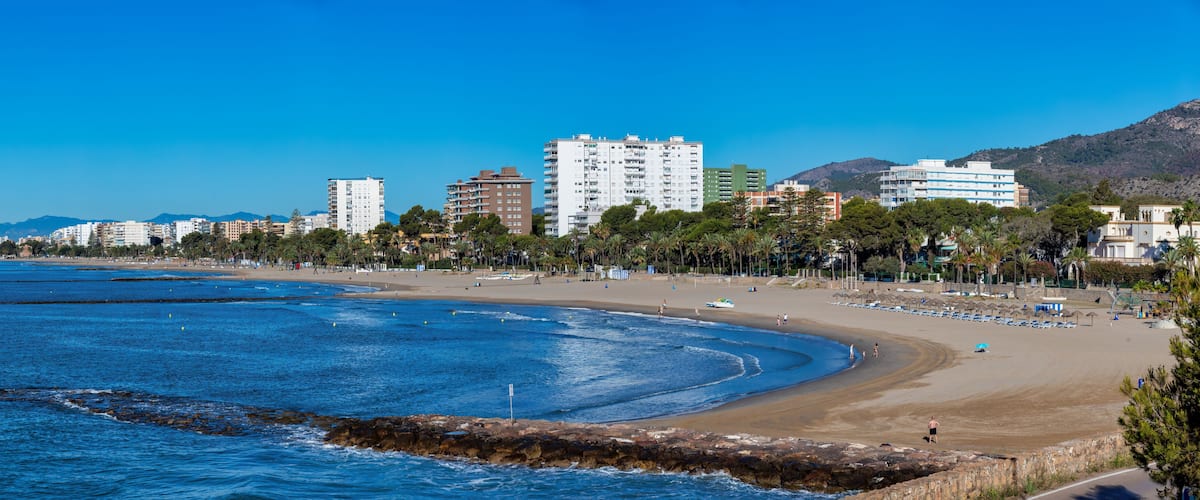 A coastal cityscape with a sandy beach, a rocky breakwater, and a backdrop of mountains.